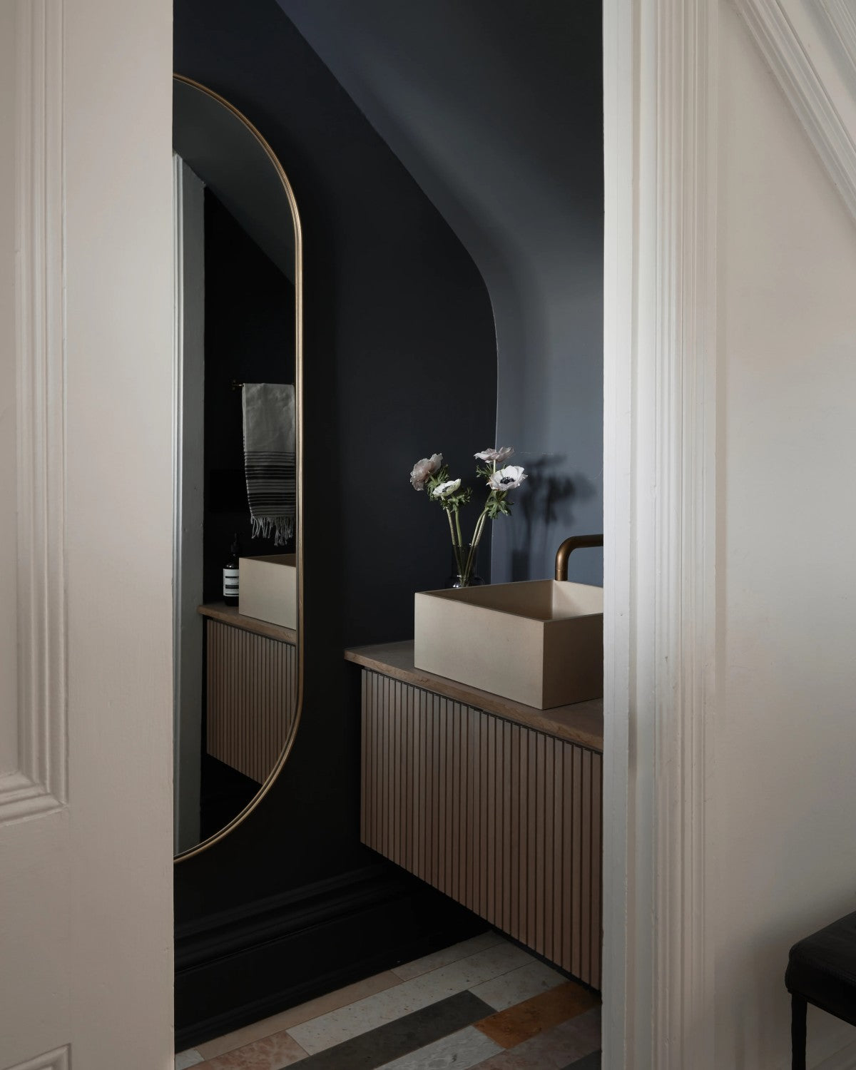 A modern bathroom with dark walls, a large oval mirror, a light wood vanity, a square sink, and a small vase with flowers on the countertop.