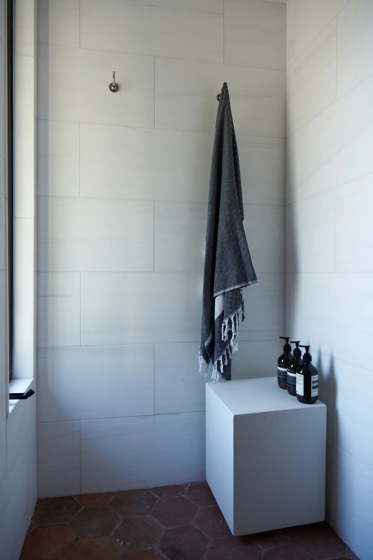 A gray towel hangs on a hook above a white cube with three bottles on top in a minimalist, tiled bathroom corner.