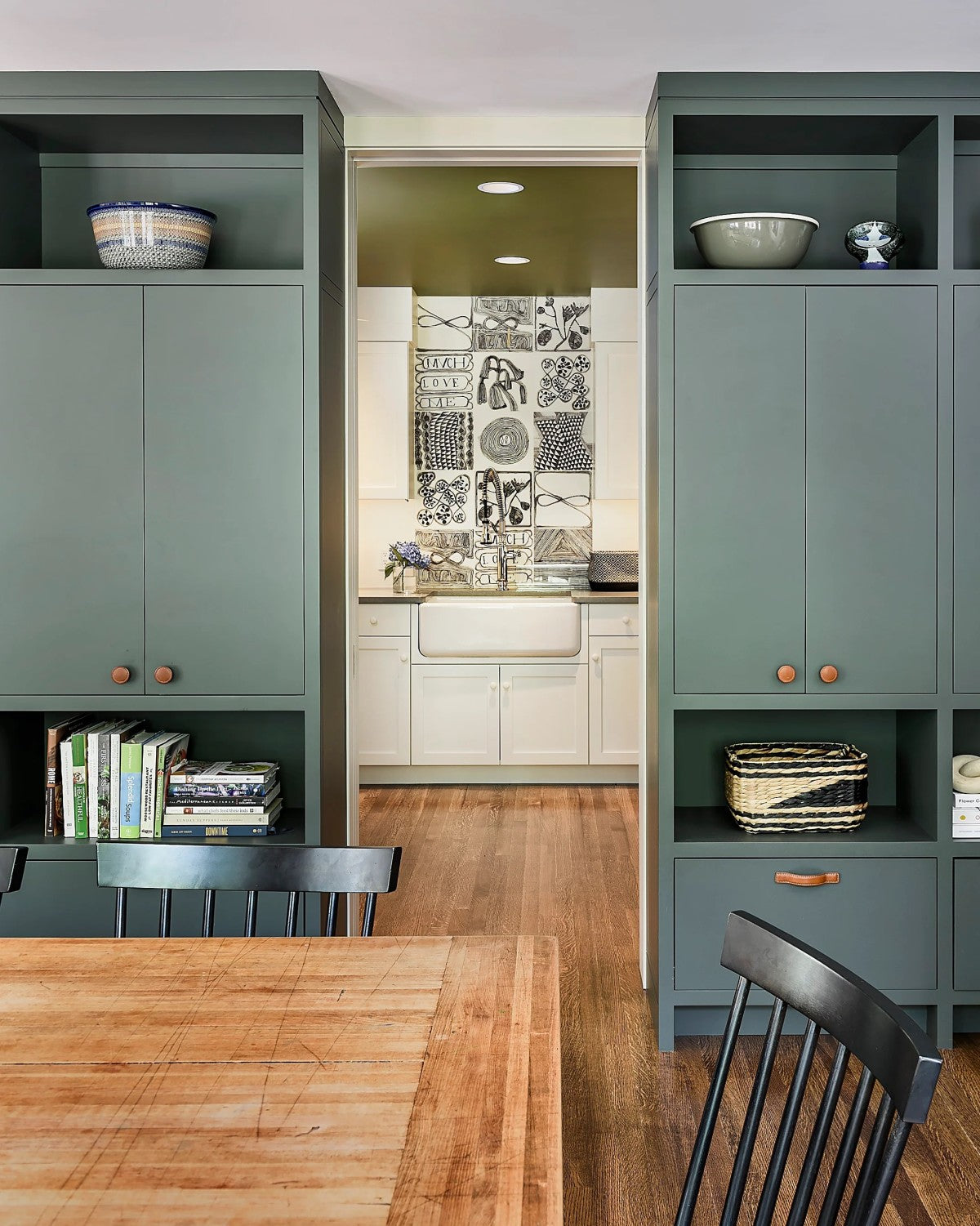 Green cabinetry frames a view into a white kitchen with a farmhouse sink and decorative black-and-white tile backsplash; wooden table and black chairs in the foreground.