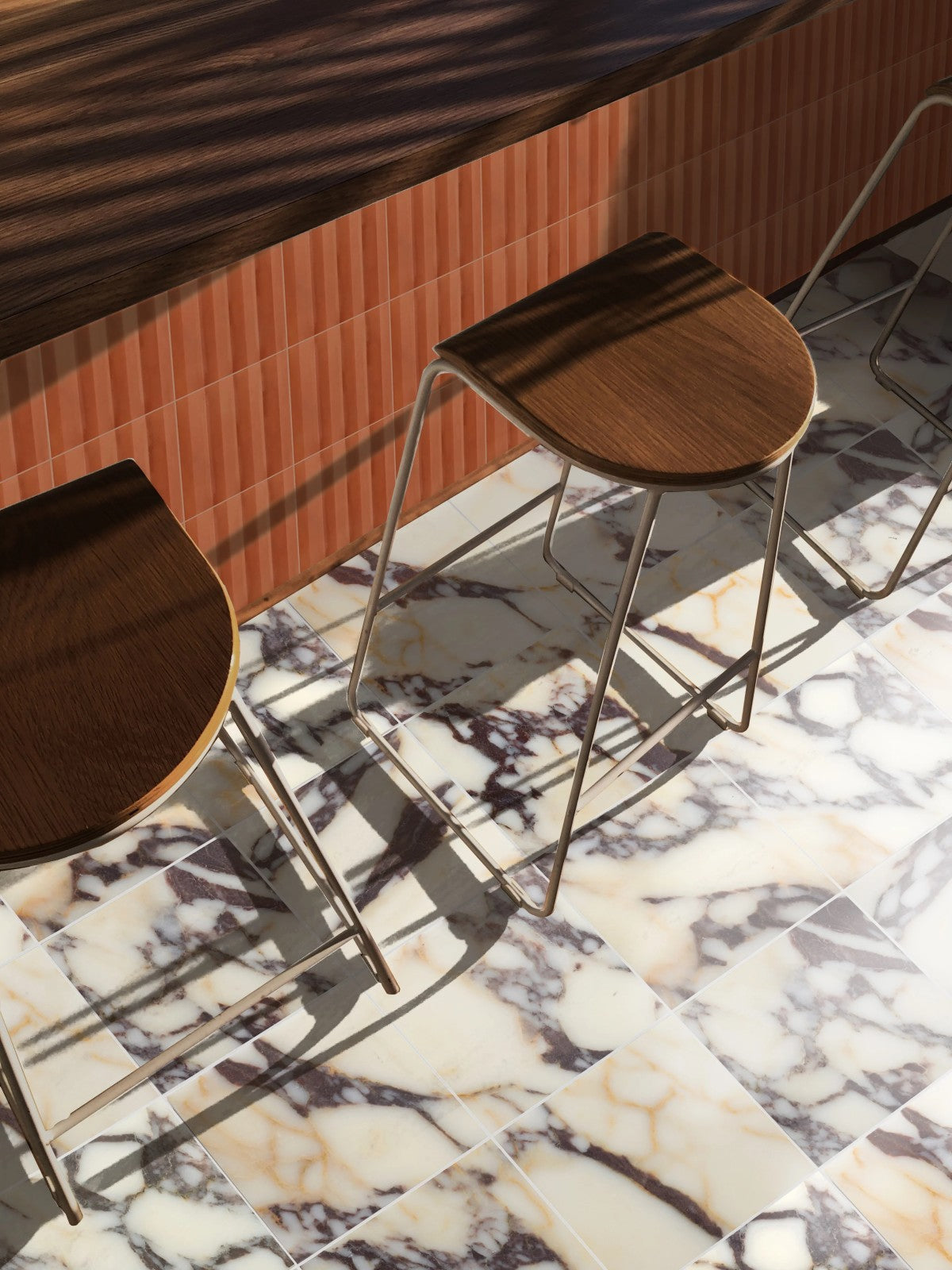 Two wooden bar stools with metal legs are placed beside a counter with a reddish-brown tiled front on a floor with a bold white and black marble pattern.