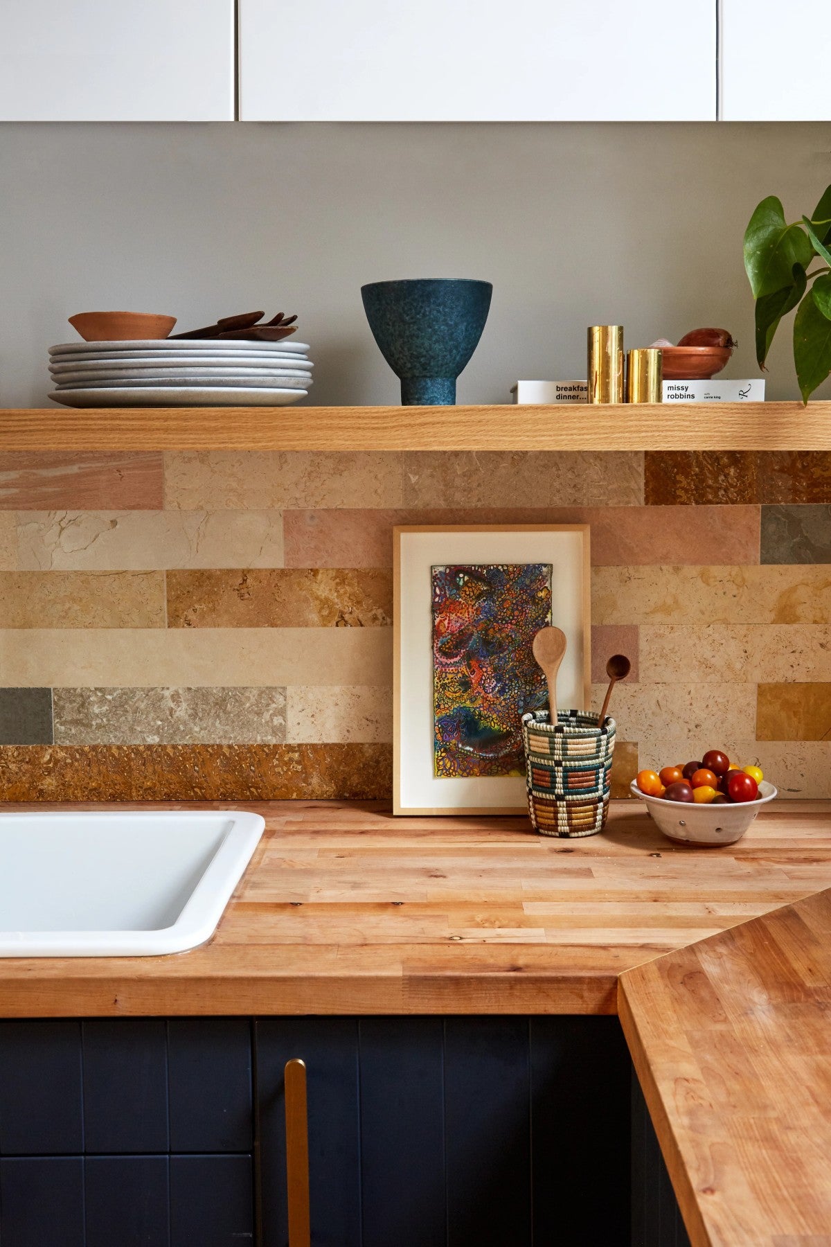 A modern kitchen with wooden countertops, an open shelf holding dishes and decor, a bowl of fruit, a framed artwork, and a plant in the corner.