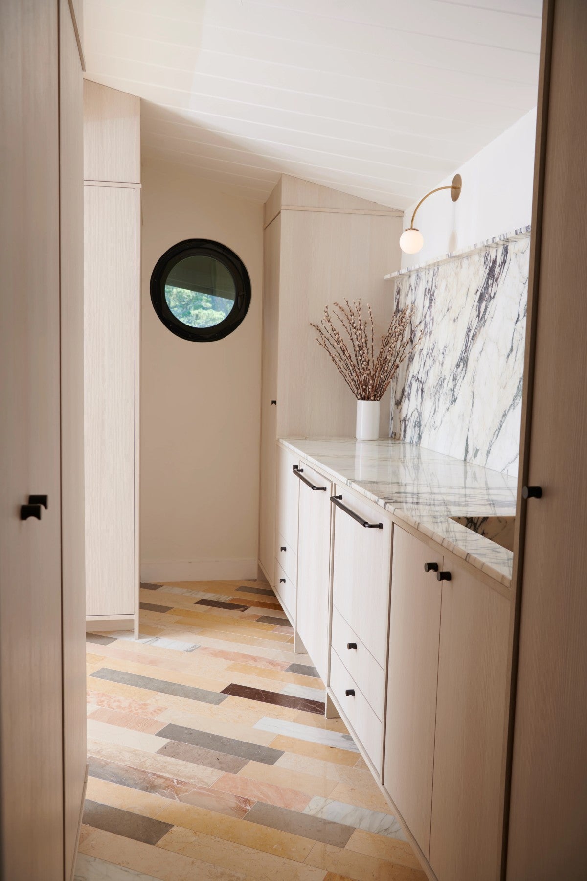 A narrow room with light wood cabinets, marble countertop and backsplash, a round window, wall sconce, and a vase with branches on the counter. The floor has a multi-toned tile pattern.
