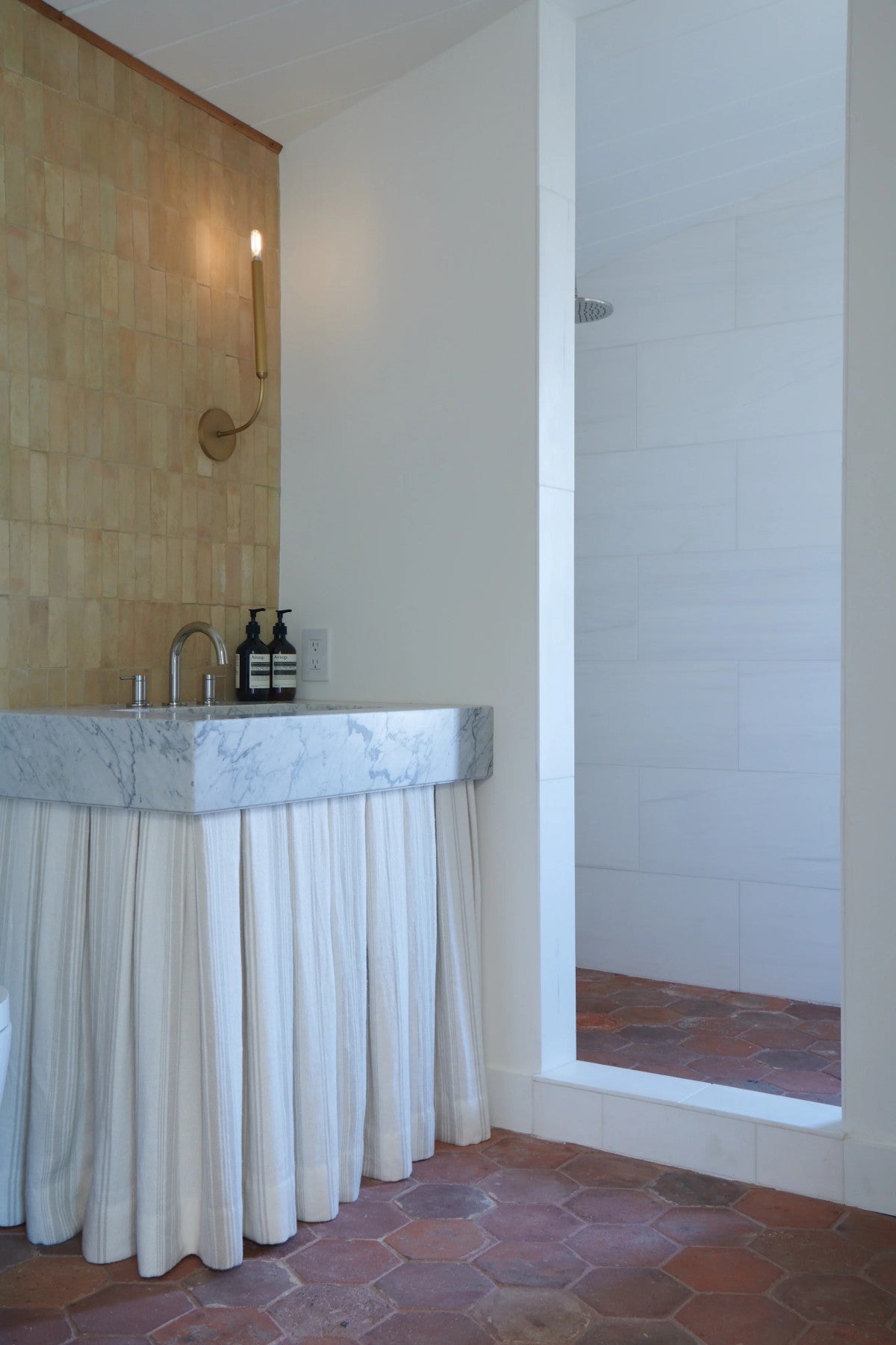 A bathroom features a marble sink with a white fabric skirt, a gold wall sconce, and an open shower with white tile and terracotta floor.