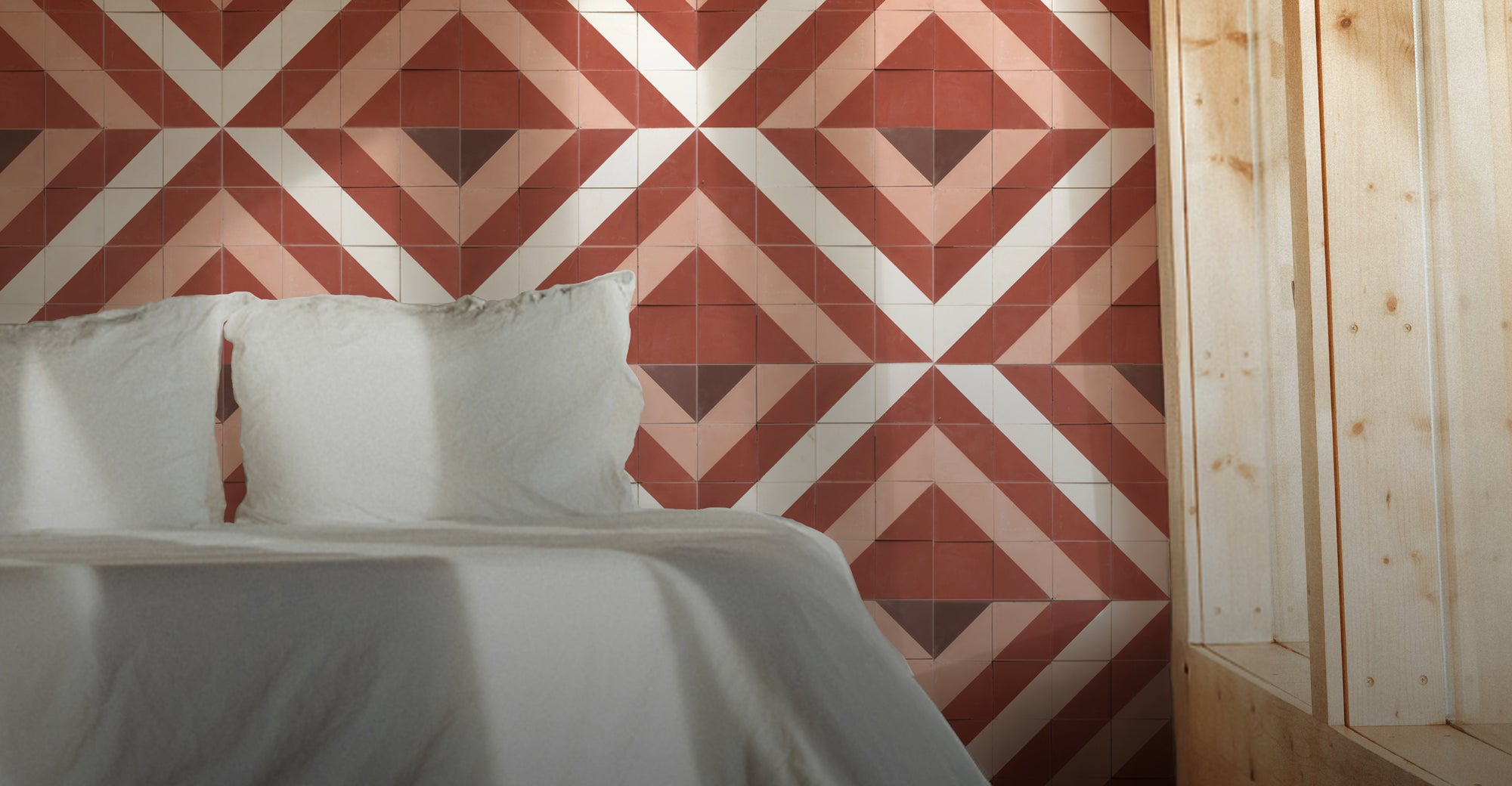 A close-up of a bed with white bedding and pillows set against a wall covered in geometric patterned tiles in terra mural red, white, and brown