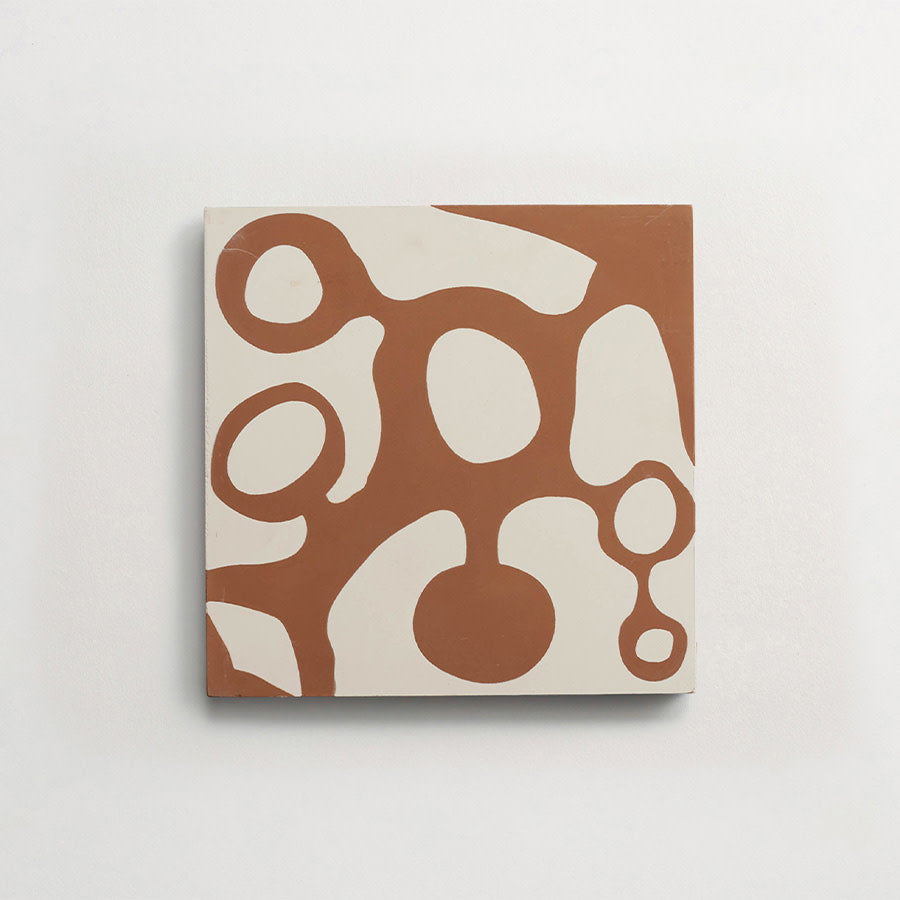 A square tile with an abstract pattern of organic, rounded brown shapes on a white background, mounted on a plain white wall.
