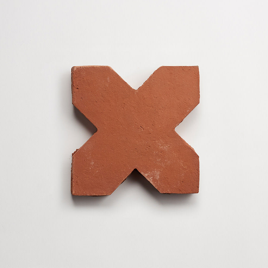A terracotta X-shaped tile with a rough texture is placed on a plain white background.