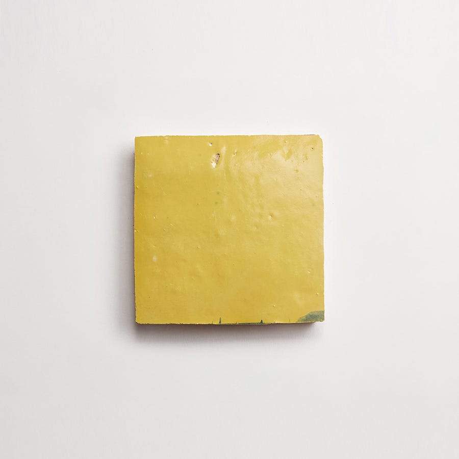 A yellow ceramic tile with a slightly uneven surface and a small chipped corner, placed on a plain white background.