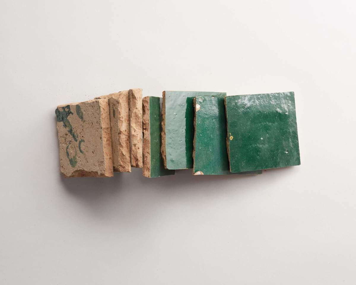 Seven square tiles are lined up on a white surface; five have green glazed tops and two are unglazed with a natural, speckled appearance.