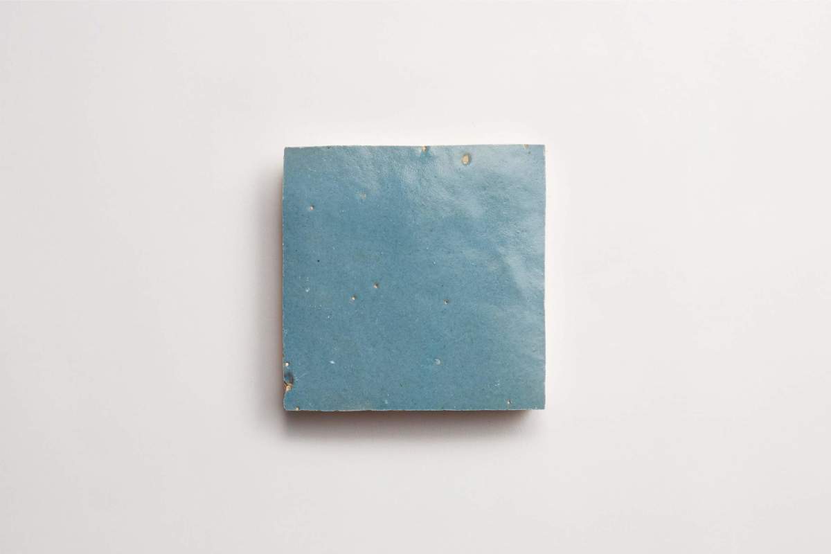 A single square blue ceramic tile with small imperfections sits on a plain white background.