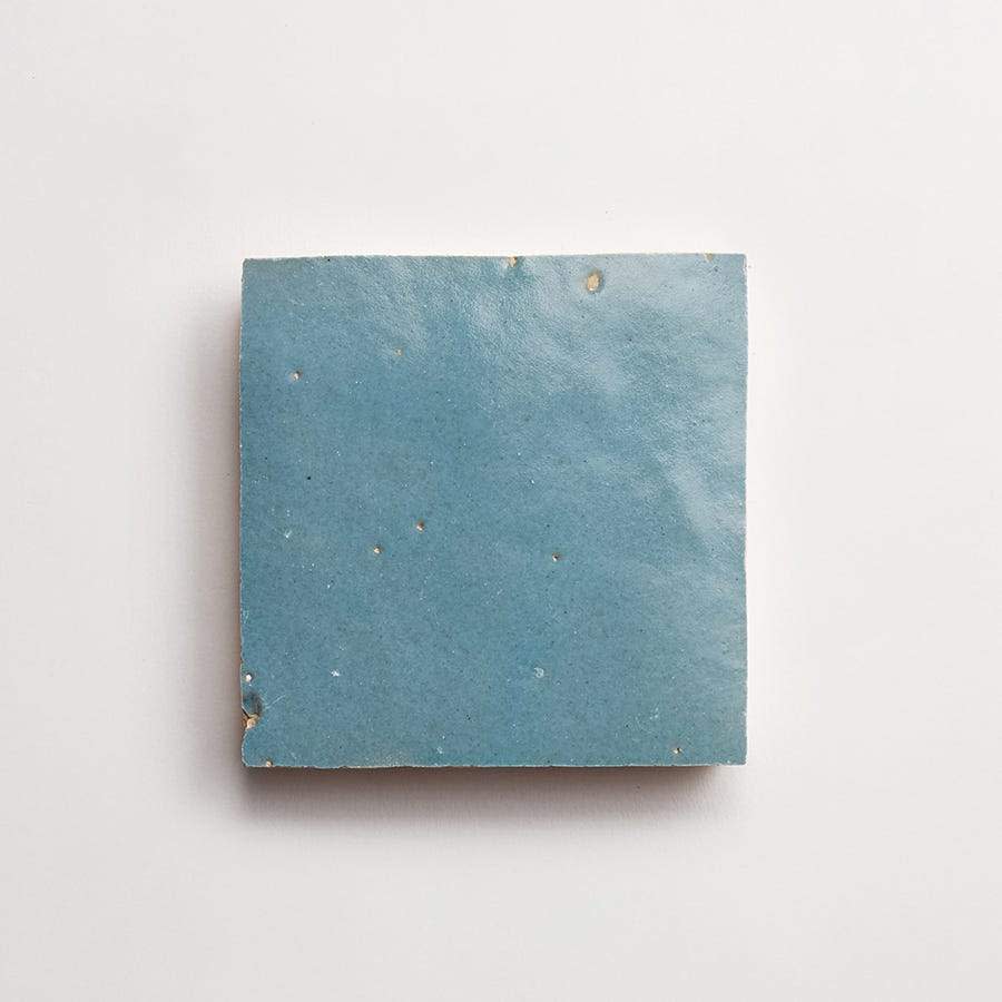 A single square ceramic tile with a smooth, matte blue surface and slight imperfections, photographed against a plain white background.