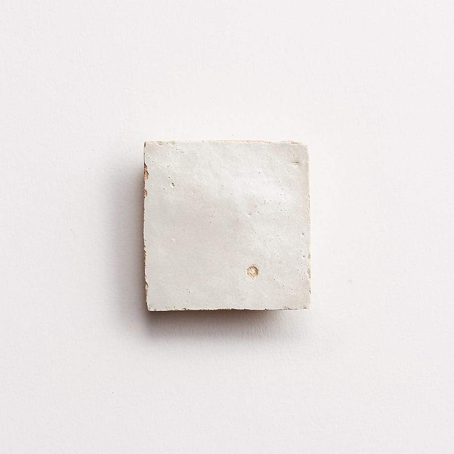 A single, square, white ceramic tile with a slightly uneven surface is centered on a plain white background.