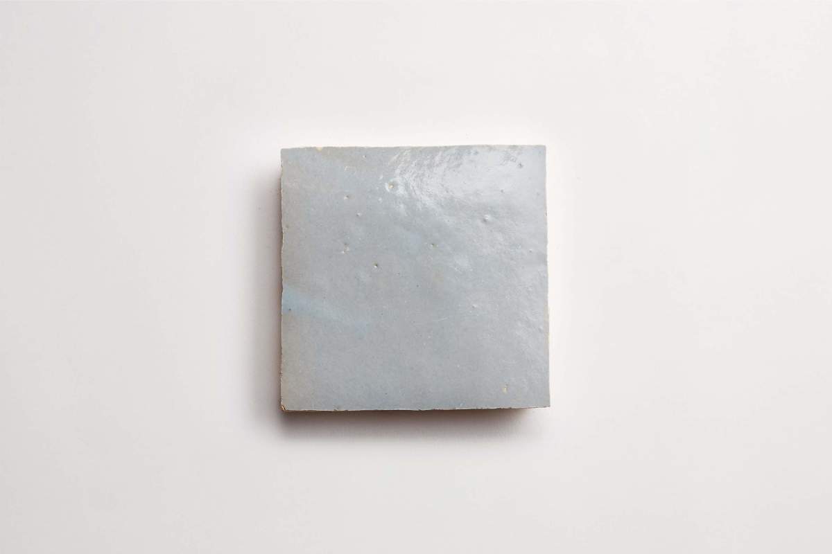 A single square light gray ceramic tile with a glossy surface and small imperfections, placed on a plain white background.