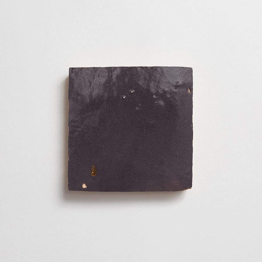 A single dark, square ceramic tile with a slightly uneven surface and small imperfections, mounted on a plain white wall.