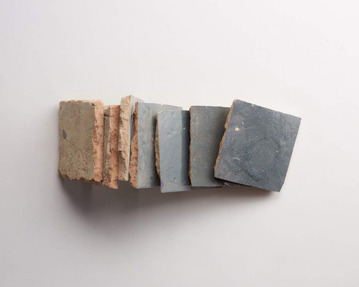 Eight square tiles with rough, beige edges and smooth, gray tops arranged in a slightly fanned-out line on a white background.