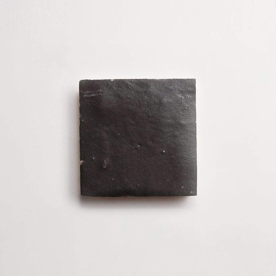 A single black square tile with a matte finish is positioned centrally on a plain white background.
