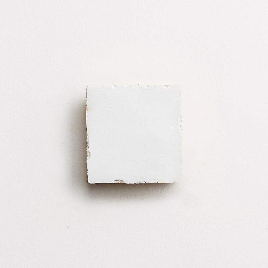 A square white tile with slightly rough edges on a light-colored background.
