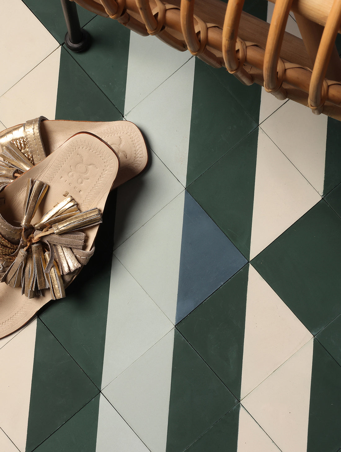 A pair of metallic gold and beige flat sandals with a large tassel detail, placed on a geometrically patterned floor of dark green, light gray, and white tiles.