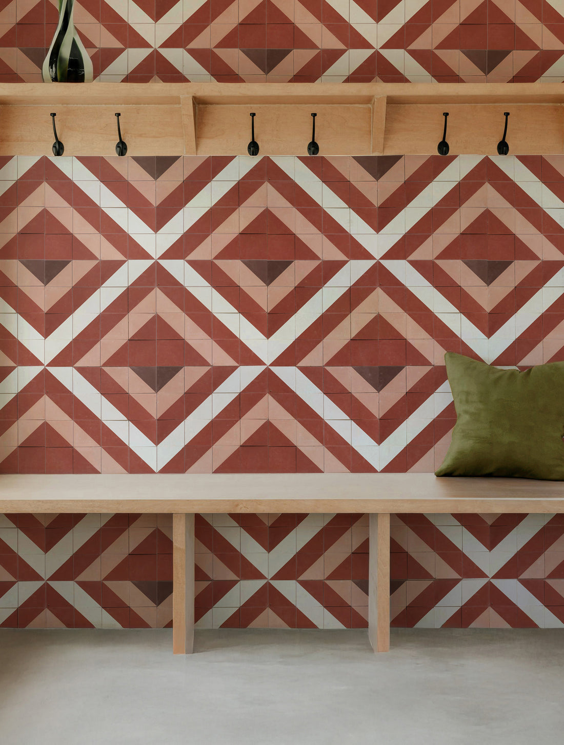 A styled interior corner featuring a geometric patterned tile wall in terracotta, beige, and white, with a light wood bench, wall hooks, and an olive green pillow.