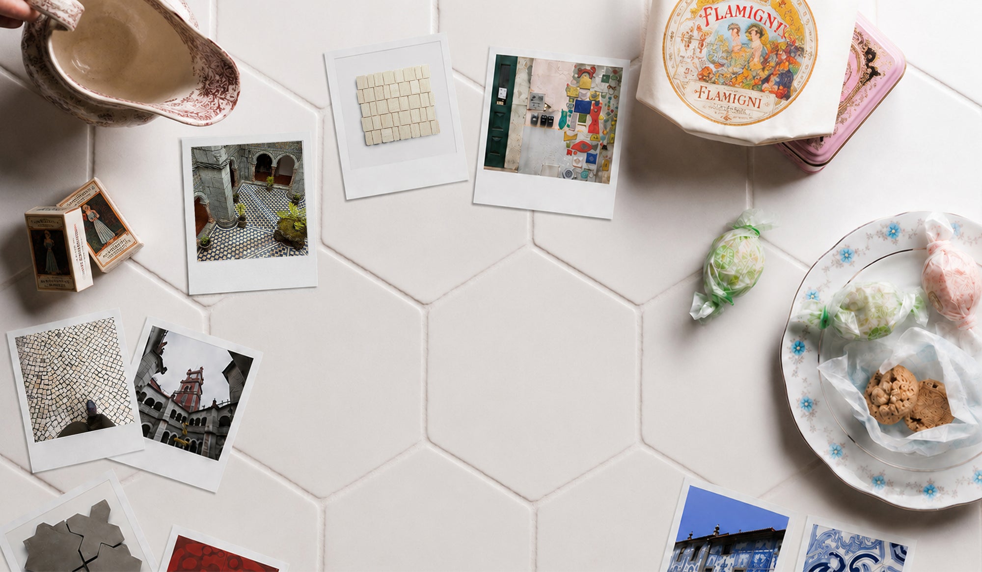 Ceramic pitcher, tin box, and plate of wrapped candies arranged on white hexagonal tiles with scattered Polaroid photos.